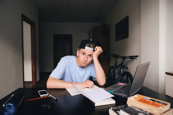 Ένας κουρασμένος μαθητής κάθεται στο γραφείο του στο δωμάτιό του. Εργασία στο σπίτι. Μια ματιά στην κάμερα. — Φωτογραφία Αρχείου
