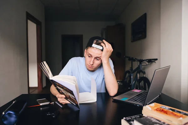 Μια θλιβερή έφηβος διαβάζει ένα βιβλίο στο γραφείο κοντά στον υπολογιστή στο δωμάτιό του. Διδασκαλία στο σπίτι. Ο φοιτητής δεν θέλει να διαβάσει το βιβλίο. — Φωτογραφία Αρχείου