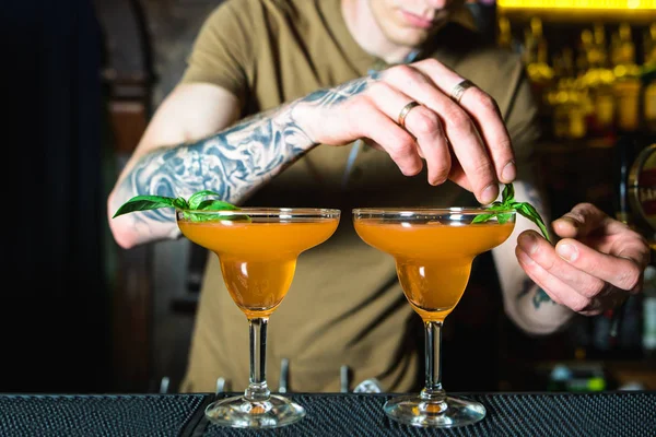 Bartender itu menciptakan minuman beralkohol oranye yang indah. Seorang bartender bertato menghiasi koktail alkohol hijau. Barman sedang mendekorasi koktail — Stok Foto