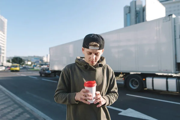 Uno studente giovane ed elegante in piedi per strada e guardando una tazza di caffè nelle sue mani. Studente sta contro un bordo della strada e camion bianco . — Foto Stock