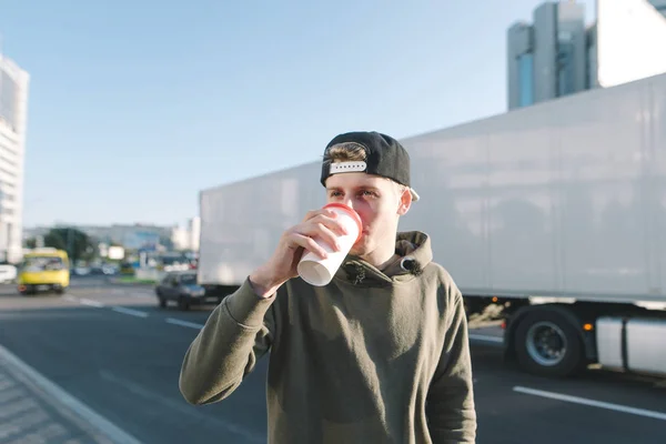 Un joven bebe café mientras camina por las calles de la ciudad. El estudiante está parado en el fondo de una carretera y un camión grande blanco . — Foto de Stock