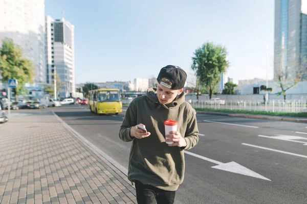 Um jovem elegante com uma xícara de café em suas mãos vai ao longo da calçada e usa um telefone celular. Um estudante escreve uma mensagem no fundo da estrada e carros . — Fotografia de Stock