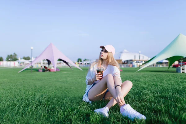 Menina bonita nova que descansa ao sentar-se em um parque de diversões na grama verde, em um dia quente com limonada em seus braços . — Fotografia de Stock