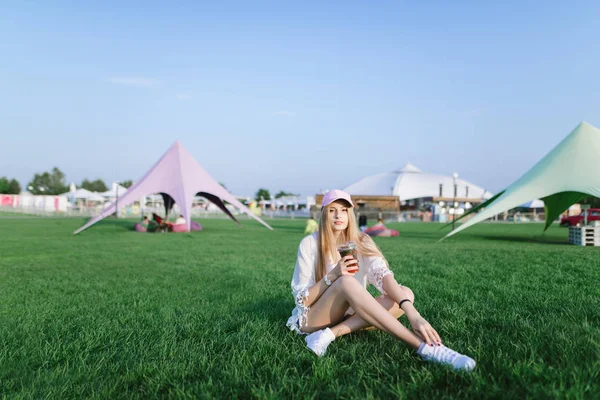 Retrato de una chica positiva que descansa en un hermoso parque en un césped verde y bebe una bebida refrescante . —  Fotos de Stock