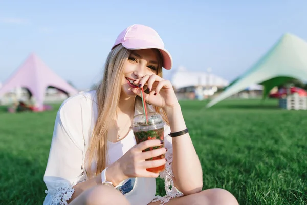 Sorrindo bela jovem bebendo um coquetel enquanto descansa em um belo parque em um gramado verde . — Fotografia de Stock