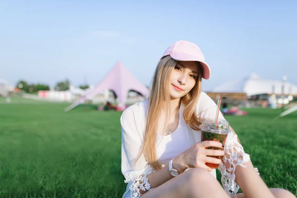 Uma jovem elegante com um boné descansando em um belo parque moderno e segurando uma limonada em suas mãos . — Fotografia de Stock