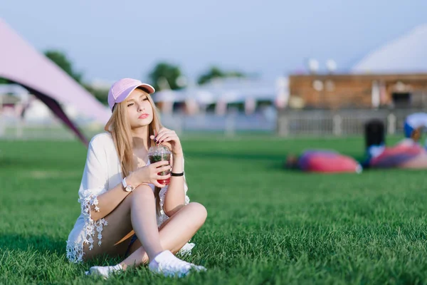 Menina elegante em um boné descansando em um gramado verde no meio de um parque de recreação, e mantém uma bebida em suas mãos . — Fotografia de Stock