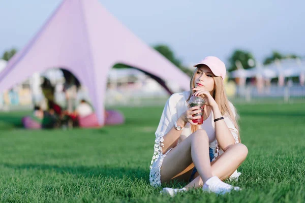 Şık kız bir serinletici kokteyl yeşil çim bir parkta oturan ve uzağa bakarak içecekler. Geri kalan kavramı.