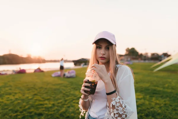 Porträt einer stilvollen jungen Frau, die mit Limonade in der Hand vor dem Hintergrund des Sonnenuntergangs steht. — Stockfoto