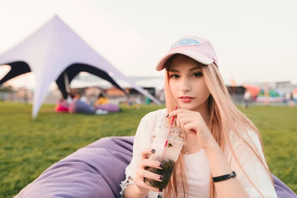 Positiv schönes Mädchen sitzt in einem Park auf einem Stuhl Tasche und hält eine Limonade in den Händen. — Stockfoto