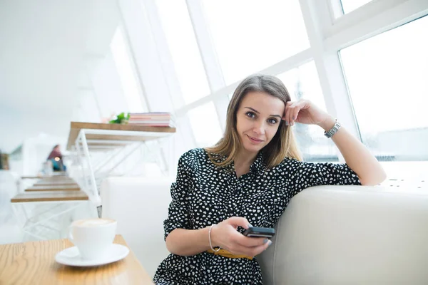 Mulher de negócios positiva com um telefone em suas mãos sentado no sofá em um elegante café moderno pela janela . — Fotografia de Stock