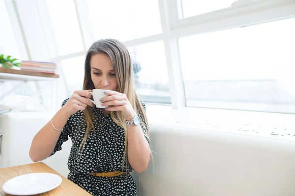 Красивая молодая женщина в кафе держит чашку чая, видна через окно со зданиями и световыми отражениями. Она отводит взгляд. Концепция образа жизни . — стоковое фото