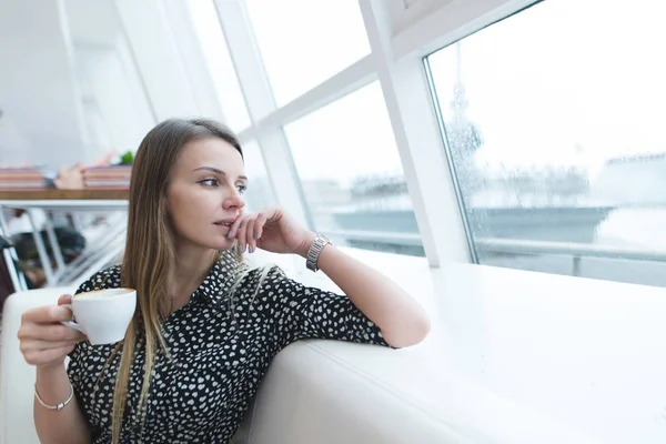 En affärskvinna med en kopp kaffe i hennes händer sitter i en modern, ljus restaurang och ser ut genom fönstret. En allvarlig kvinna ser ut i fönstret och tycker. — Stockfoto