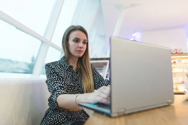 Retrato de una hermosa mujer de negocios sentada en un café de diseño moderno y trabajando en una computadora portátil. Estilo de vida. Una mirada enfocada a la computadora . —  Fotos de Stock