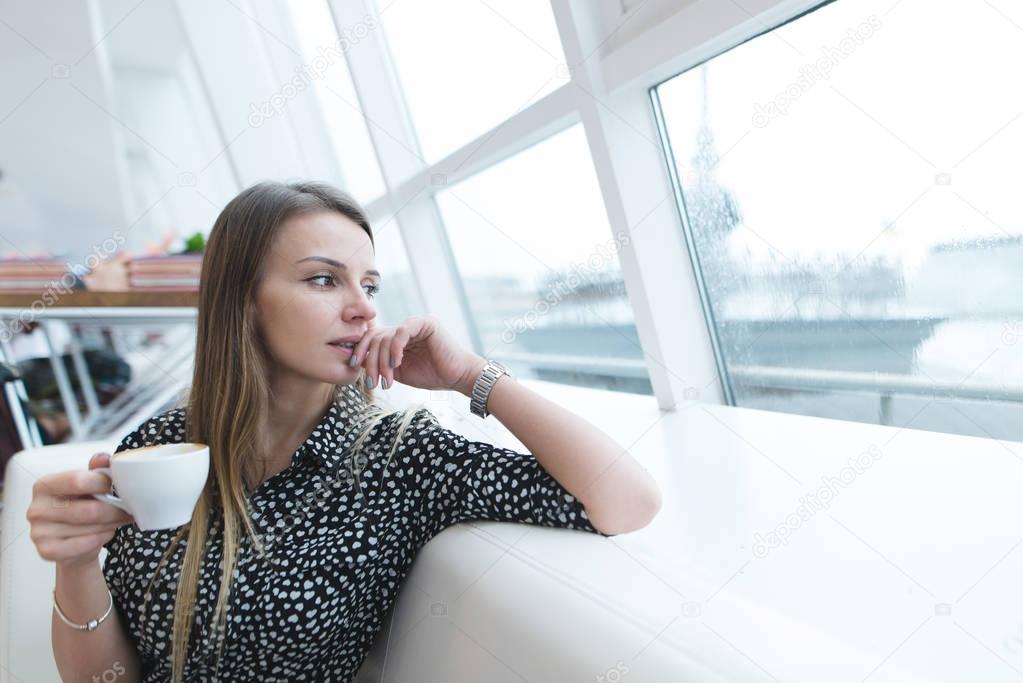 A business woman with a cup of coffee in her hands sits in a modern, light restaurant and looks out the window. A serious woman looks in the window and thinks.