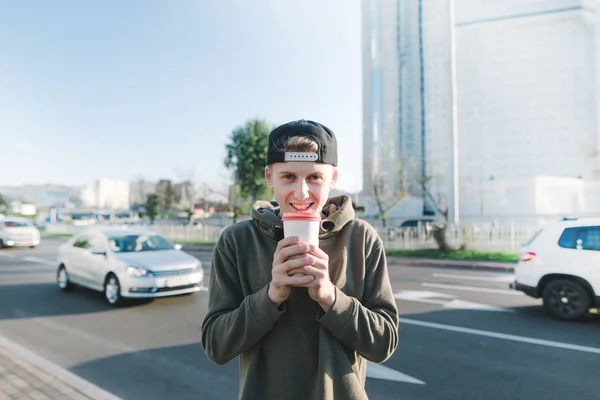 Un joven positivo está de pie junto a la carretera con coches con una taza de bebida caliente y sonrisas. Estudiante con café en el fondo de la ciudad — Foto de Stock