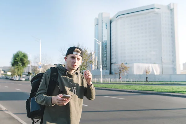 Jovem elegante com uma mochila andando pela rua e ouvindo música em fones de ouvido contra o pano de fundo da cidade. Estilo de vida e conceito de pessoas . — Fotografia de Stock