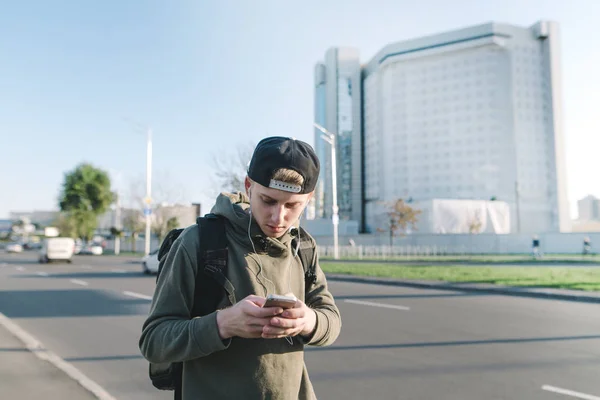 A stylish young student walking down the street with headphones in his ears and looking at his own snapphone on the background of the city. — Stock Photo, Image