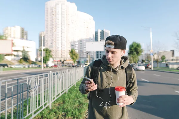 A young man with headphones in the ears and coffee in the yoke strolls around the city and uses the phone. Lifestyle and people concept. Lifestyle and people concept.
