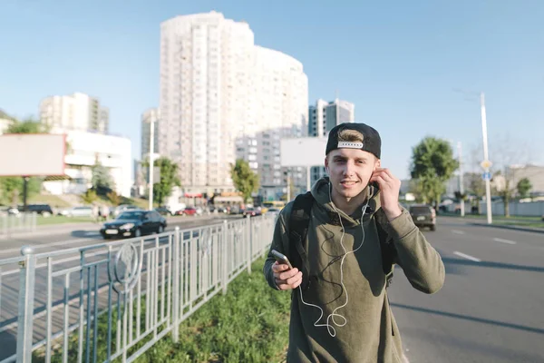 Красивый улыбающийся молодой человек со смартфоном в руках и наушниками в ушах наслаждается музыкой во время прогулки по городу. На фоне улицы . — стоковое фото
