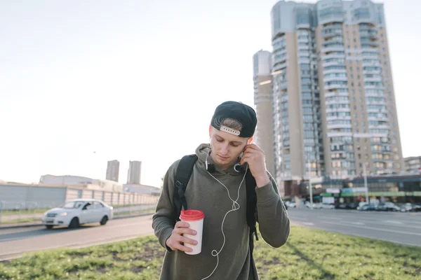 Un hombre guapo con un vaso de bebida caliente en sus manos escucha música y corrige los auriculares en sus oídos contra el telón de fondo del paisaje urbano. Estilos de vida — Foto de Stock