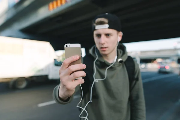 Das Telefon steht im Fokus auf dem Hintergrund eines verschwommenen jungen Mannes mit Kopfhörern und urbaner Landschaft. der Student schaltet die Musik am Telefon ein. Lebensstil und Menschen-Konzept. — Stockfoto