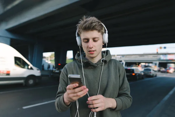Um jovem estudante ouvindo música e escrevendo uma mensagem no telefone aguardando transporte público. No fundo da ponte e da estrada. Estilo de vida e conceito de pessoas . — Fotografia de Stock