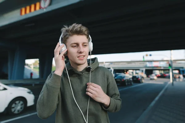 Un estudiante positivo escuchará música en los auriculares mientras espera el transporte público en una parada de autobús debajo del puente. Concepto de estilo de vida y personas . — Foto de Stock