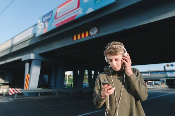 Уличный портрет молодого человека, который пишет сообщение на смартфоне и слушает музыку. Молодой человек ждет общественного транспорта на автобусной остановке под мостом . — стоковое фото