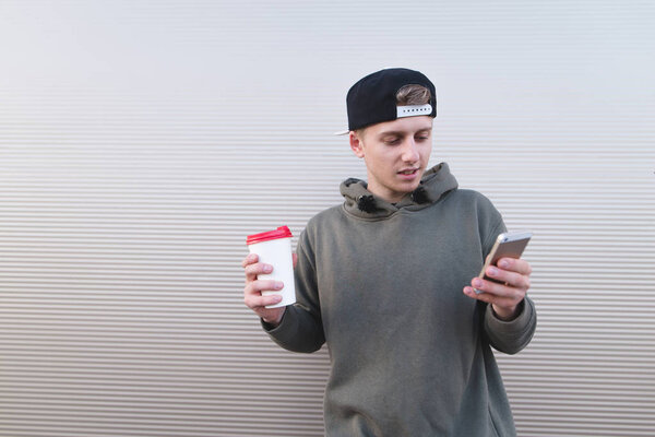 A young man stands on the background of a light wall with a cup of beverage in his hand and uses a smartphone.