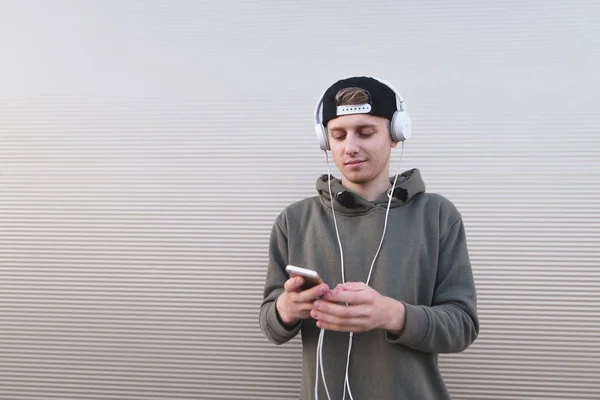 Um jovem ouve música nos fones de ouvido e olha para seu smartphone no fundo de uma parede de luz . — Fotografia de Stock