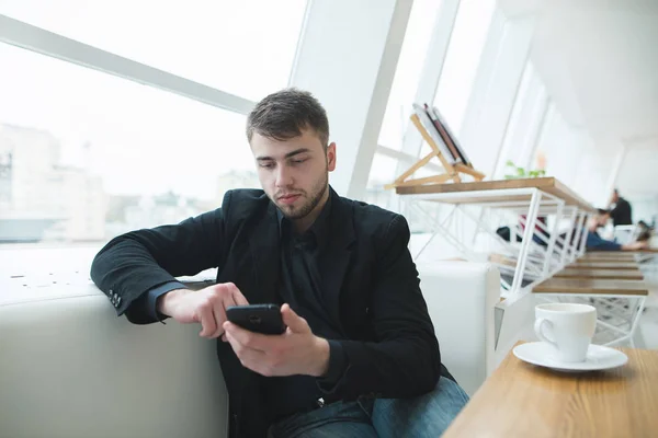 Fiatal üzletember ül egy étteremben ablak egy telefonnal a kezében. Egy ember ír egy üzenetet egy kávézóban, és a modern design. Kávészünet. — Stock Fotó