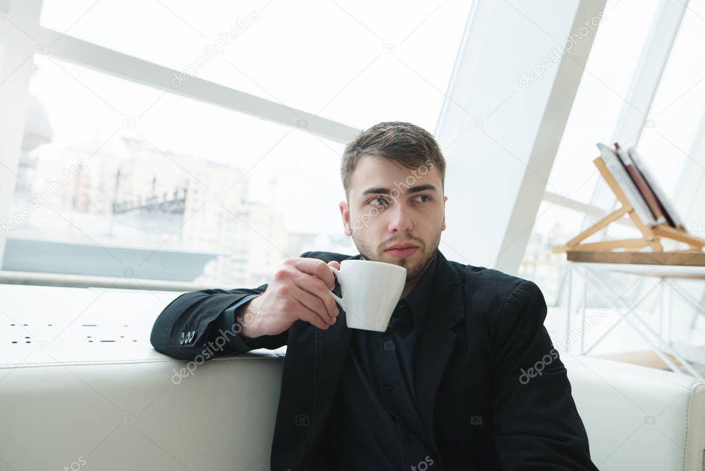 A man with a beard sits on a beautiful light cafe with a cup of coffee in his hands. Coffee break in the restaurant.