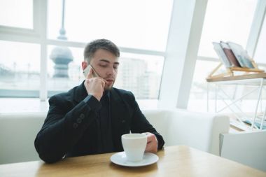 Bir kafede bir fincan için bir kafede telefonla konuşmak. Takım elbiseli bir adam şık bir restoranda pencerenin yakınındaki masada oturur ve telefonda iletişim kurar