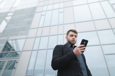 Kentsel modern mimari zemin karşı daha düşük bir açıdan elbiseli bir adam sokak portresi. Ciddi bir genç adam yürürken sakallı telefonu kullanıyor.