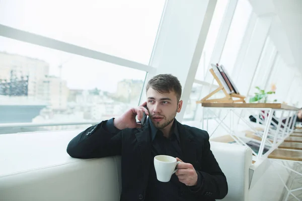 Портрет чоловіка, який розмовляє по телефону з чашкою гарячого напою в руках. Чоловік з бородою сидить у стильній кав'ярні та обідає. Перерва на каву . — стокове фото