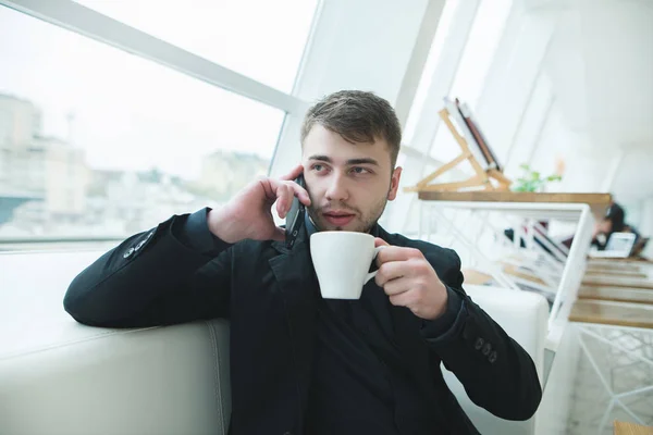 Мужчина сидит у окна и разговаривает по телефону с чашкой горячего кофе в руках. Обед в стильном кафе с современным дизайном. Перерыв на кофе . — стоковое фото
