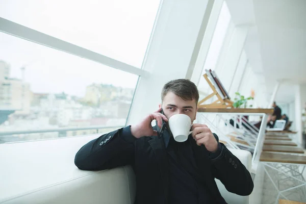 Un hombre de negocios guapo se sienta en un restaurante moderno y habla por teléfono. Pausa para almorzar café. Un hombre bebe café de una taza . —  Fotos de Stock