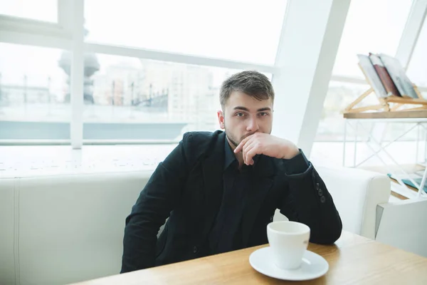 Portret poważne mężczyzny w garniturze, który siedzi w kawiarni, z filiżanką kawy i patrzy w kamerę. Biznesmen siedzi w jasnej restauracji w pobliżu okna podczas lunchu. Rzut oka na kamery. — Zdjęcie stockowe