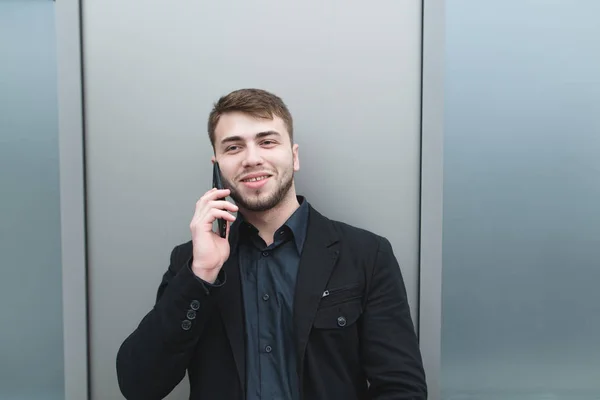 Un hombre positivo con traje y barba habla por teléfono en el fondo de una pared metálica y sonríe. Retrato de negocios . —  Fotos de Stock