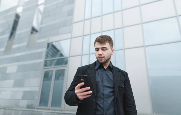 Business portrait in the open air. A businessman writes a message on a smartphone. On the background of routine architecture. — Stock Photo, Image