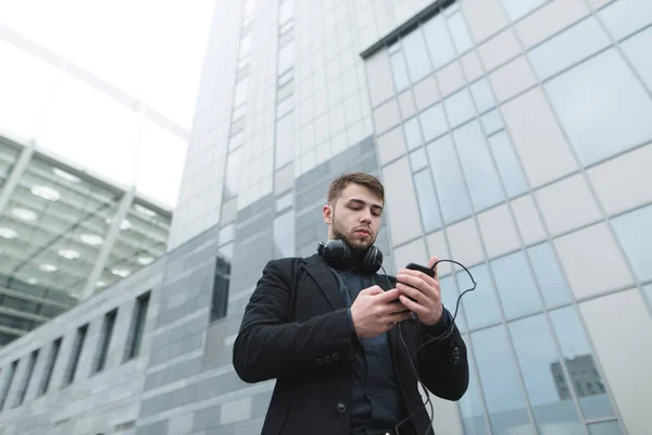Уличный портрет бизнесмена с бородой, который смотрит на свой смартфон и собирается слушать музыку из наушников, которые у него на шее . — стоковое фото
