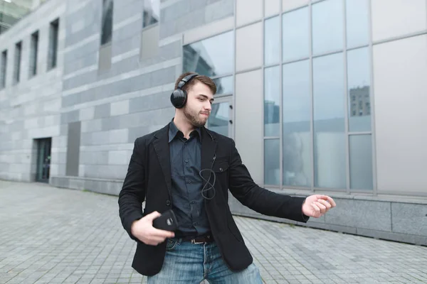 Позитивный человек в куртке танцует под музыку в наушниках на фоне городского пейзажа. Радостный позитивный бизнесмен рад . — стоковое фото