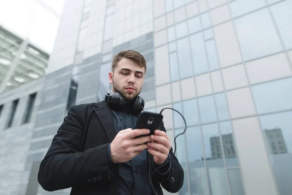 Уличный портрет серьезного человека с наушниками на шее, выбирающего музыку на телефоне на фоне городского пейзажа . — стоковое фото
