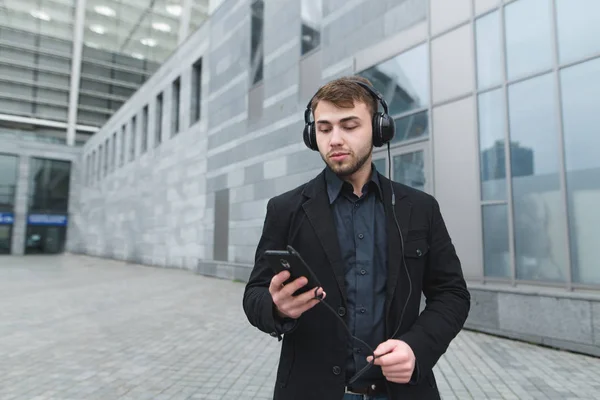 Вуличний портрет це серйозно чоловік у костюмі, який слухає музику в навушниках і використовує Інтернет на телефоні під час прогулянки містом . — стокове фото