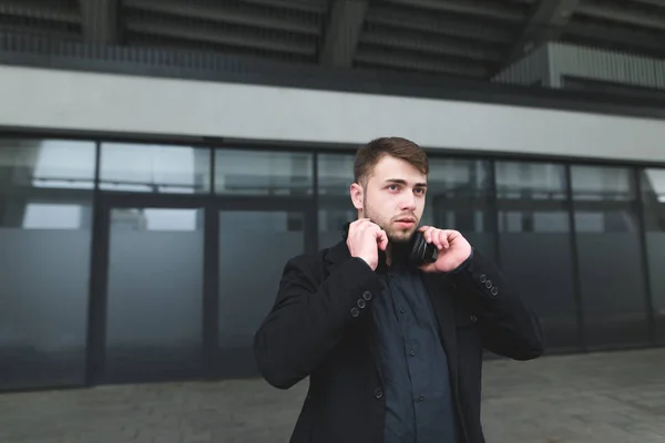 Un joven hombre de negocios con barba y auriculares en el cuello posa sobre el fondo de las paredes de un edificio moderno. Un hombre de traje se encuentra en el fondo de la arquitectura moderna . — Foto de Stock