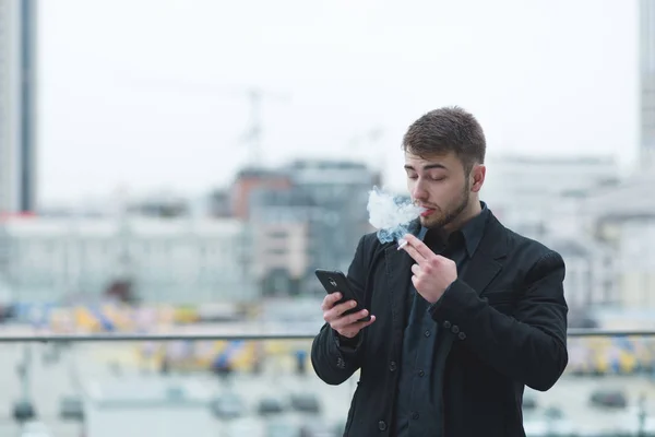A young businessman with a beard smokes a cigarette on a snack. A man smokes a cigarette in the street against the background of the city and looks at the smartphone.