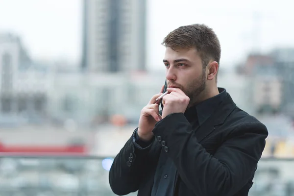 Um homem de negócios fuma um cigarro e fala por telefone no fundo de uma paisagem da cidade . — Fotografia de Stock