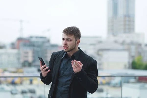An emotional businessman with a telephone in his hands smokes a cigarette and is angry with the background of a city-blurred landscape. An angry man calms himself with cigarettes. — Stock Photo, Image