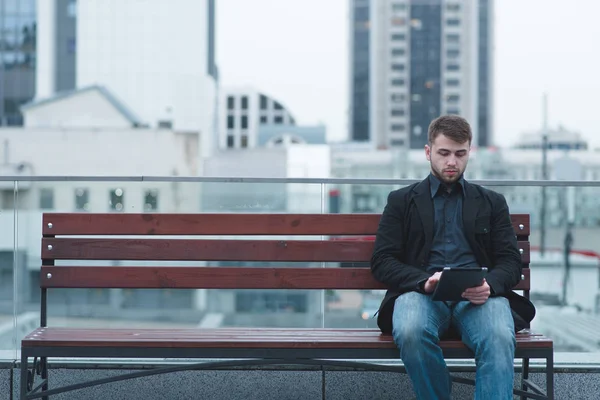 Der Mann sitzt auf einer Bank im Hintergrund einer begrünten Wiese und bedient sich eines Tablets. ein Mann ruht mit einem Gerät auf einer Bank. — Stockfoto